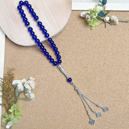 Blue beaded prayer beads (masbaha) with silver tassel on a beige surface with white flowers and green leaves.